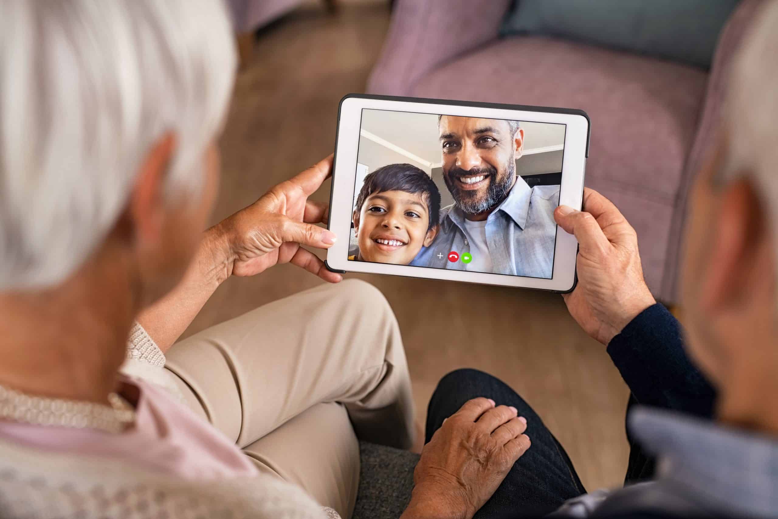 Rear view of senior couple doing a video call with son and grandson during covid19 pandemic. Close up of hands of old grandparents holding digital tablet while in conversation with little grandson. Happy indian father with child greeting in video conference his parents.