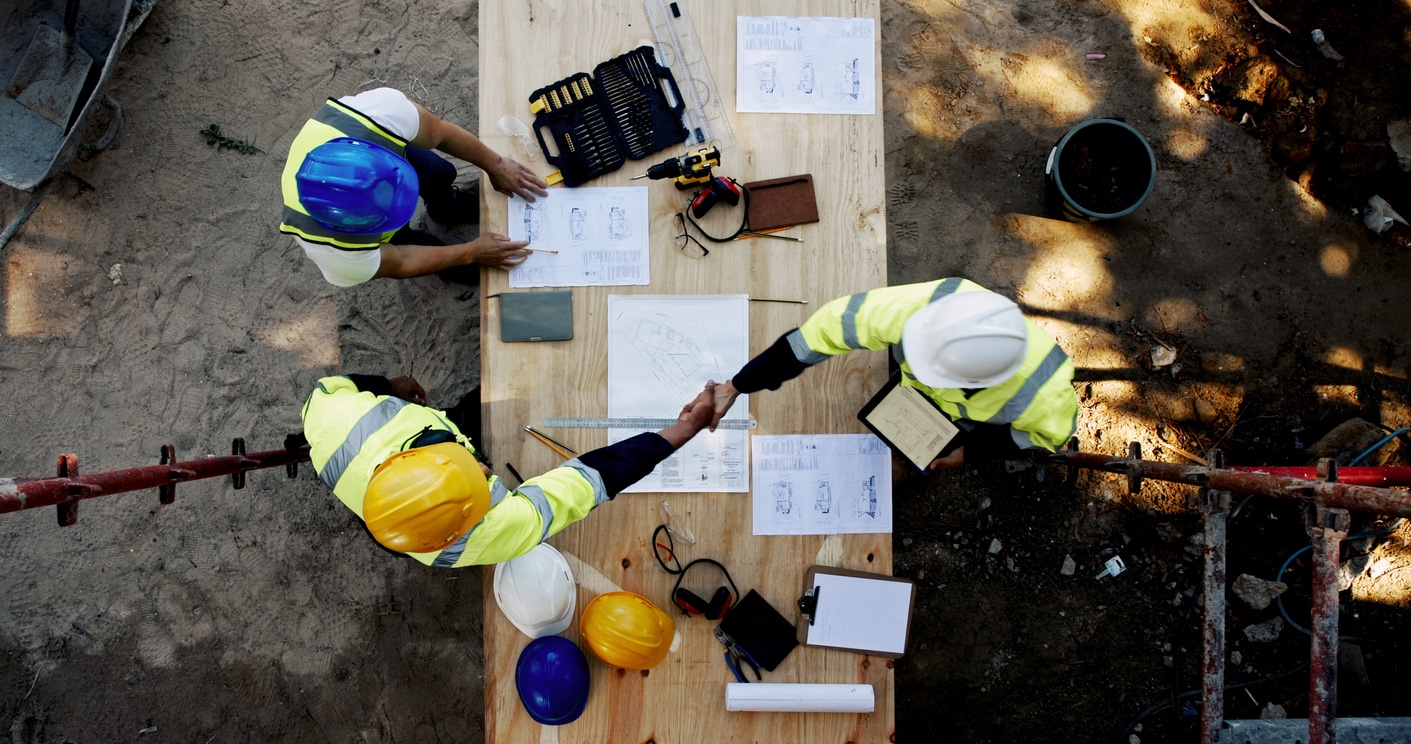 Blueprint, architecture and men at table shaking hands for collaboration, agreement and inspection from above\. Construction, building team and contractor on site with floor plan, handshake and deal