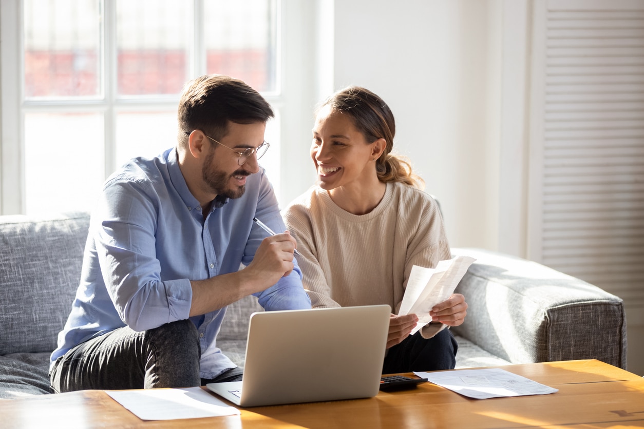Happy young married couple sit on couch calculate expenses use easy online banking service at home, smiling millennial husband and wife count taxes house expenditures pay bills on internet