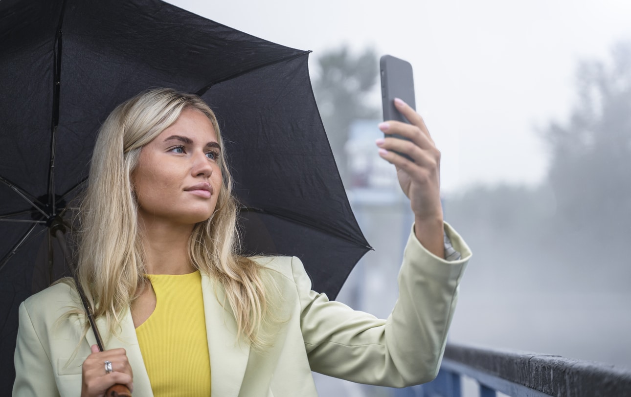 Portrait of an attractive young woman standing under an umbrella during the rain and taking a selfie alone during the day, natural blue and gray background\.