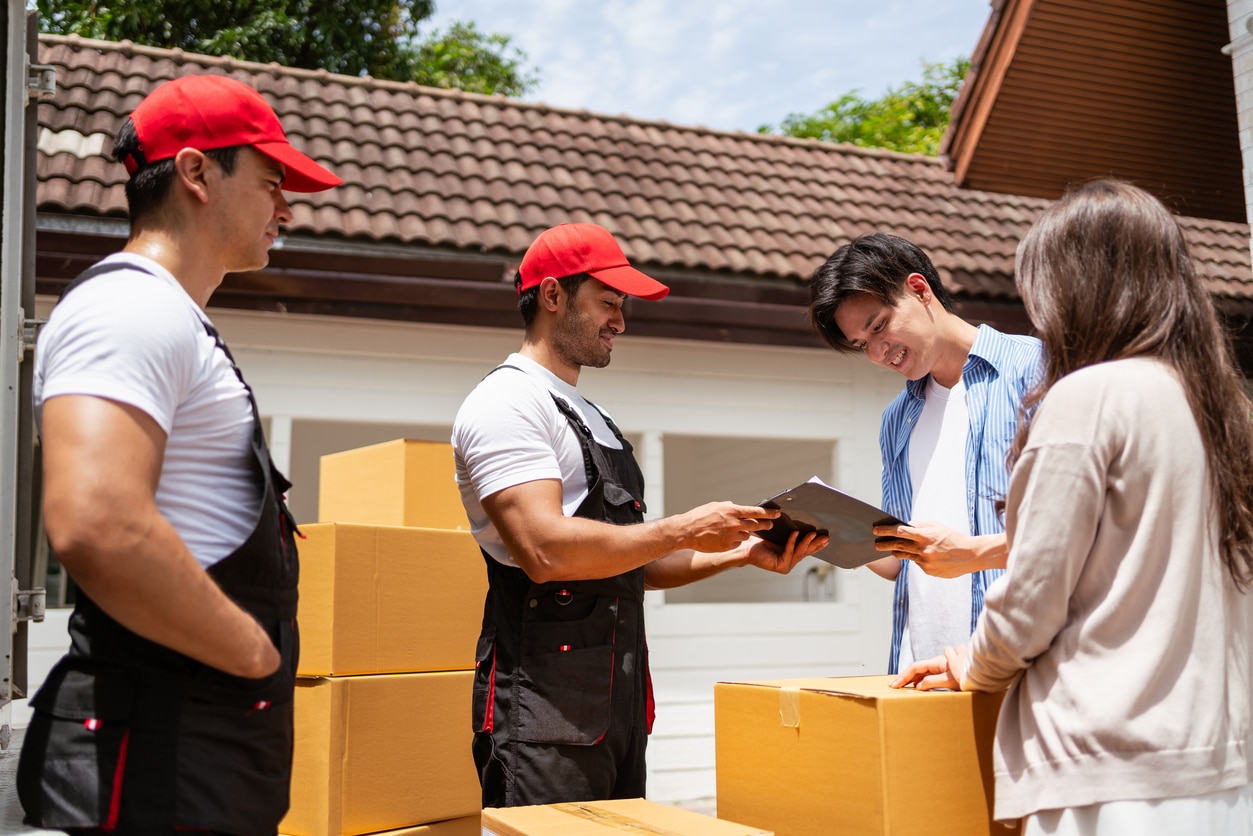 A couple signs the checklist of Asian and Caucasian workers in uniform unloading cardboard boxes from the truck\. Delivery men unloading boxes to couple home\. Professional delivery and moving service\.