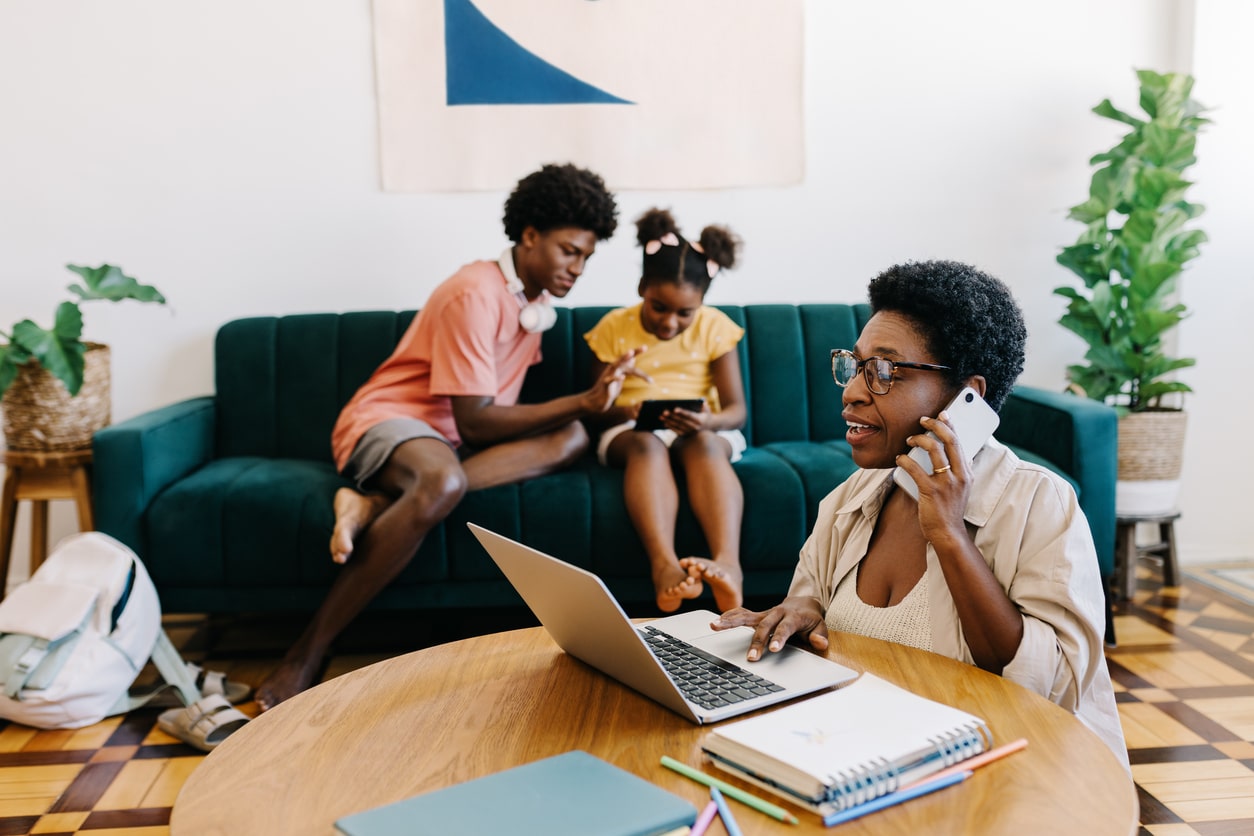 Woman working on a laptop and making a mobile phone call while her kids sit on a couch in the background\. Working mom communicating with her colleagues from her home office\.