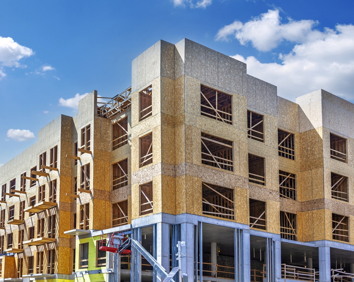 Multi\-story residential building under construction using wood frame and OSB sheathing on a concrete podium base\.